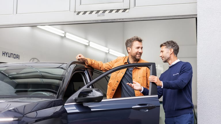 Ein Serviceberater im Hyundai-Polohemd bespricht mit einem Kunden ein Fahrzeug an der geöffneten Fahrertür in der Serviceannahme.