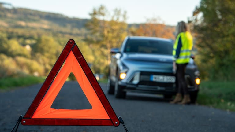 Warndreieck auf der Straße, dahinter ein Hyundai am Straßenrand und eine Fahrerin in Warnweste