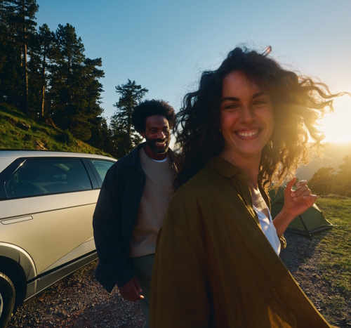 Eine Frau und ein Mann lächeln im Freien neben einem Auto und einem Zelt bei Sonnenuntergang, mit Bäumen im Hintergrund.